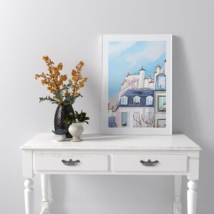 May include: A white console table with two drawers and turned legs. The table is decorated with a black vase with orange berries and a small potted plant. A framed print of a watercolor painting of a Parisian building with a blue sky is hanging on the wall above the table.