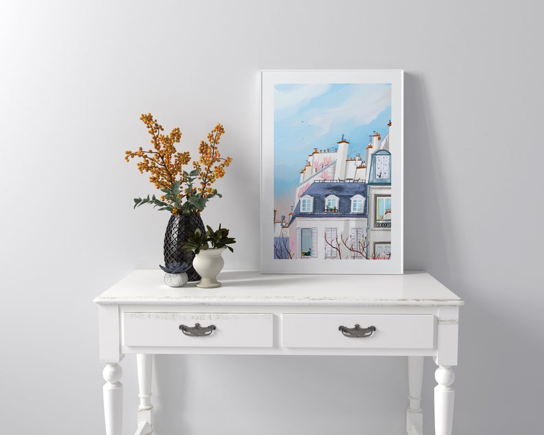May include: A white console table with two drawers and turned legs. The table is decorated with a black vase with orange berries and a small potted plant. A framed print of a watercolor painting of a Parisian building with a blue sky is hanging on the wall above the table.