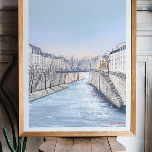 Peut inclure: Une peinture encadr&eacute;e d'un paysage urbain parisien avec une rivi&egrave;re au premier plan. La rivi&egrave;re est de couleur bleu clair et les b&acirc;timents sont blancs avec un ciel bleu clair en arri&egrave;re-plan. La peinture est sign&eacute;e "Lynda Bakker" en bas &agrave; droite.