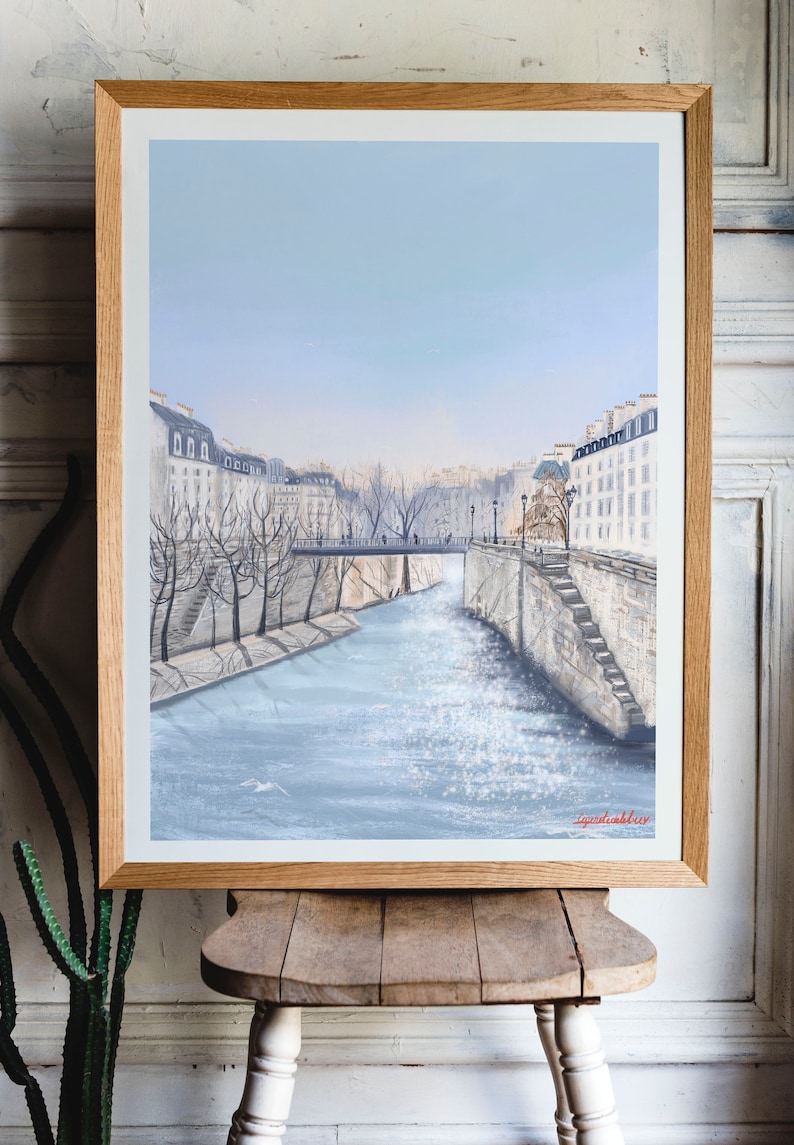 Peut inclure: Une peinture encadr&eacute;e d'un paysage urbain parisien avec une rivi&egrave;re au premier plan. La rivi&egrave;re est de couleur bleu clair et les b&acirc;timents sont blancs avec un ciel bleu clair en arri&egrave;re-plan. La peinture est sign&eacute;e "Lynda Bakker" en bas &agrave; droite.
