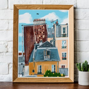 May include: A framed painting depicting a colourful cityscape with multiple buildings. The buildings are painted in shades of brown, yellow, blue, and grey. The painting has a blue sky with white clouds and a small bird flying in the sky.