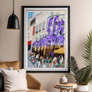 May include: Framed art print depicting a Parisian cafe scene. The artwork features a building with a red "HOTEL" sign, purple wisteria, and people seated at tables under a yellow awning. The print is framed in black with a gold inner border.