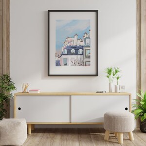 May include: A framed watercolor painting of a Parisian building with a blue roof and white walls. The building has several windows and a balcony. The painting is set against a light blue sky with white clouds.