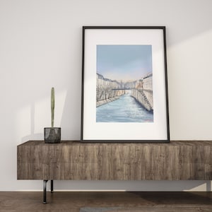 Peut inclure: Une peinture &agrave; l'aquarelle en noir et blanc encadr&eacute;e repr&eacute;sentant une sc&egrave;ne de rivi&egrave;re dans une ville. La peinture repr&eacute;sente une rivi&egrave;re avec des b&acirc;timents de chaque c&ocirc;t&eacute; et un pont au loin. Le ciel est d'un bleu clair.