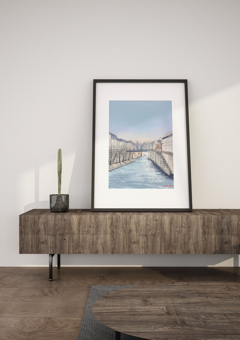 Peut inclure: Une peinture &agrave; l'aquarelle en noir et blanc encadr&eacute;e repr&eacute;sentant une sc&egrave;ne de rivi&egrave;re dans une ville. La peinture repr&eacute;sente une rivi&egrave;re avec des b&acirc;timents de chaque c&ocirc;t&eacute; et un pont au loin. Le ciel est d'un bleu clair.
