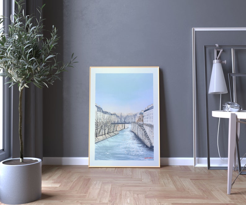 Peut inclure: Une peinture encadr&eacute;e repr&eacute;sentant une ville avec une rivi&egrave;re qui la traverse. Les b&acirc;timents sont blancs et la rivi&egrave;re est bleue. Le ciel est bleu clair. La peinture est dans un cadre dor&eacute;.