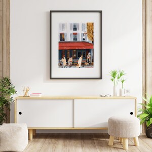 May include: A framed illustration of a Parisian street scene with a cafe called "Paris" and people walking by. The cafe has a red awning and a white facade with windows. The illustration is in a black frame and is hanging on a white wall.