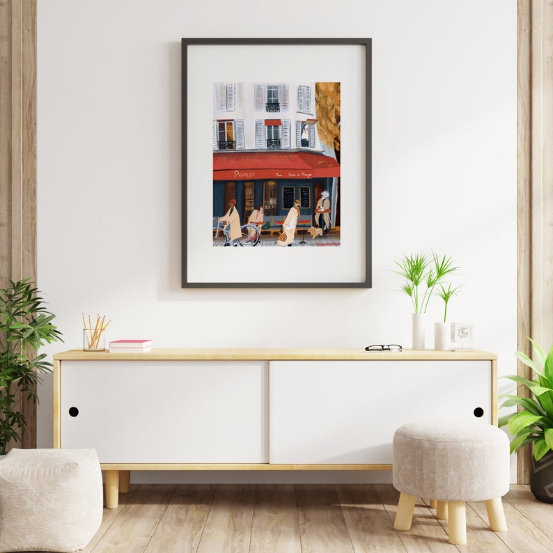 May include: A framed illustration of a Parisian street scene with a cafe called "Paris" and people walking by. The cafe has a red awning and a white facade with windows. The illustration is in a black frame and is hanging on a white wall.