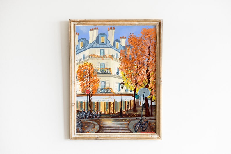 May include: A framed painting depicting a Parisian street scene with a white building, fall foliage, and bicycles parked in front of a cafe. The cafe has a sign that reads "Boulangere".