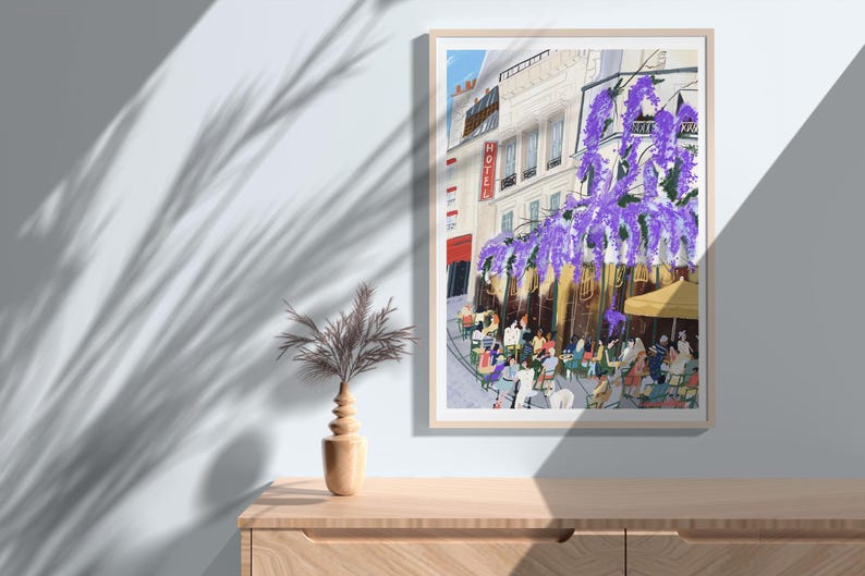 May include: A framed art print depicts a Parisian cafe scene. The artwork showcases a building with a "HOTEL" sign, purple wisteria, and people seated at tables. The print is displayed on a wooden cabinet, with a decorative vase and plant.