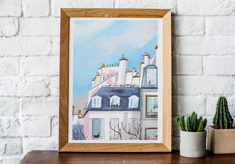May include: A framed watercolor painting of a Parisian cityscape with a blue sky and pink flowers on the rooftops. The buildings are white with gray slate roofs and windows.