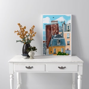 May include: A framed illustration of a Parisian cityscape with multiple buildings, including a yellow building with a blue roof and a red brick building with a grey roof. The buildings are painted in a cartoon style with a blue sky and white clouds in the background.