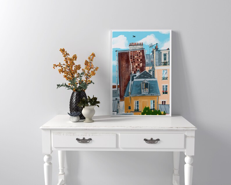 May include: A framed illustration of a Parisian cityscape with multiple buildings, including a yellow building with a blue roof and a red brick building with a grey roof. The buildings are painted in a cartoon style with a blue sky and white clouds in the background.
