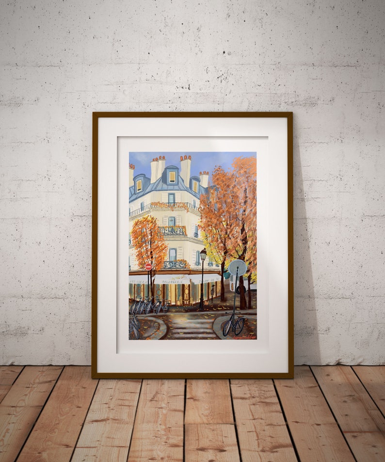 May include: A framed painting of a Parisian street scene with a cafe, bicycles, and a building with a blue roof. The trees are in autumn colors, and the sky is blue.