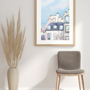 May include: A framed watercolor painting of a Parisian cityscape with white buildings and pink flowers. The painting is hanging on a white wall with a brown chair and a vase of dried grass in front of it.