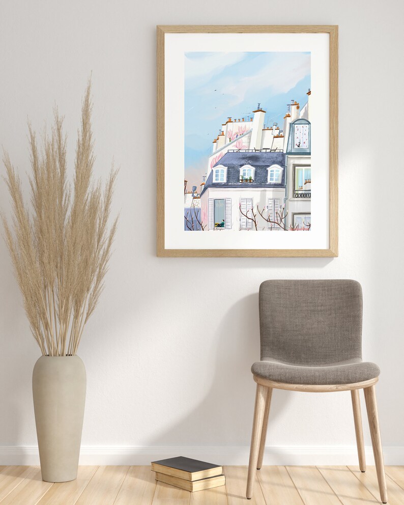 May include: A framed watercolor painting of a Parisian cityscape with white buildings and pink flowers. The painting is hanging on a white wall with a brown chair and a vase of dried grass in front of it.