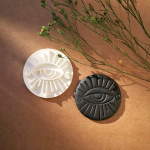 Op de afbeelding: Twee ronde, oogvormige decoratieve stukken. Eén is wit, de andere zwart. Beide hebben een oogontwerp met zonnestralen en manen. Gedroogd groen op de achtergrond, tegen een bruine ondergrond.