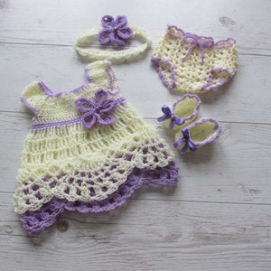 May include: A crocheted baby outfit set in yellow and purple. The set includes a dress, a headband, a pair of shoes, and a nappy cover.