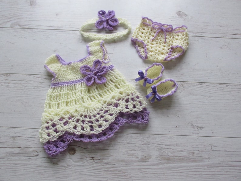 May include: A crocheted baby outfit set in yellow and purple. The set includes a dress, a headband, a pair of shoes, and a nappy cover.