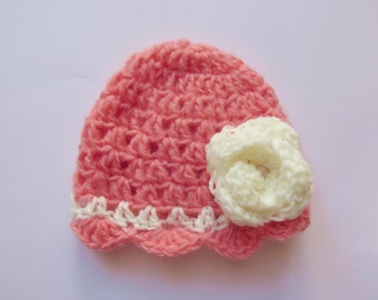 May include: A pink crocheted hat with a white crocheted flower on the side. The hat has a scalloped edge.