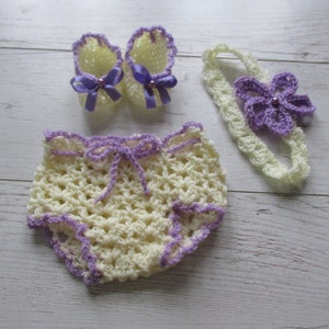 May include: A set of crocheted baby clothes in yellow and purple. The set includes a headband with a flower, a pair of booties, and a pair of shorts.