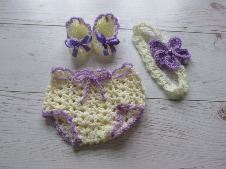 May include: A set of crocheted baby clothes in yellow and purple. The set includes a headband with a flower, a pair of booties, and a pair of shorts.