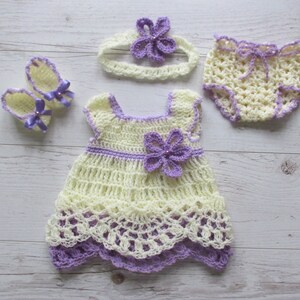 May include: A crocheted baby outfit set in yellow and purple. The set includes a dress, a headband, a pair of booties, and a diaper cover.