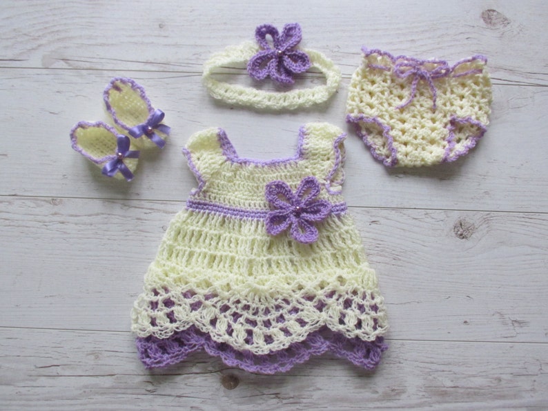 May include: A crocheted baby outfit set in yellow and purple. The set includes a dress, a headband, a pair of booties, and a diaper cover.