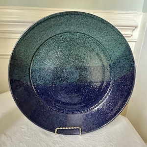 May include: A large, round ceramic plate with a speckled, gradient design. The plate transitions from a deep blue at the bottom to a teal green at the top. The plate is displayed on a gold-colored metal stand.