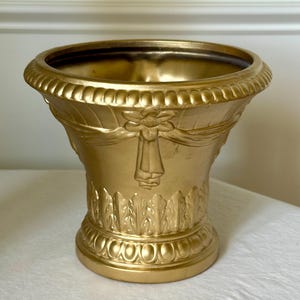 May include: A gold-colored decorative urn with a wide, open top and a detailed, ornate design. The urn features a floral motif with ribbon accents and a textured base. The overall shape is reminiscent of classical architecture.