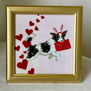 May include: A framed print featuring a black and white dog carrying a red envelope, adorned with a red headband and hearts. The artwork is set against a pink background, framed in gold.