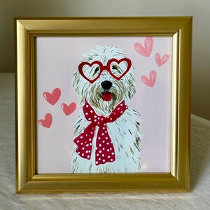 May include: A framed painting of a white dog wearing heart-shaped red glasses and a red polka-dot scarf. Pink hearts float around the dog on a pink background. The frame is gold.