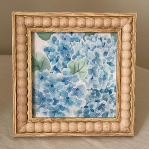 May include: Square wooden picture frame with a light brown finish. The frame has a beaded edge and contains a watercolor painting of blue hydrangea flowers and green leaves.