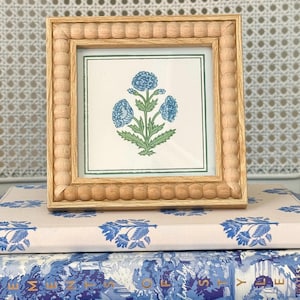 May include: A square wooden picture frame with a beaded texture border containing a print of a blue flower with green leaves on a white background. The frame is light-toned wood and features a simple, clean design with a thin green border.