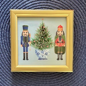 May include: A framed print featuring a Christmas tree in a blue and white vase, flanked by two nutcracker figures. The nutcrackers are dressed in blue and red uniforms. The frame is gold.