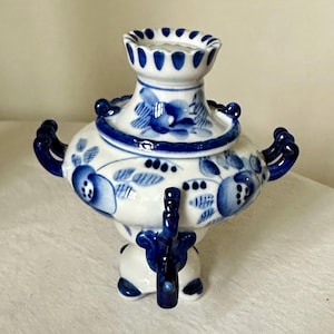 May include: A white and blue ceramic samovar with floral patterns. The samovar has a round body, a spout, and a lid with a decorative top. The handles and details are in a deep blue color. A traditional Russian tea kettle.