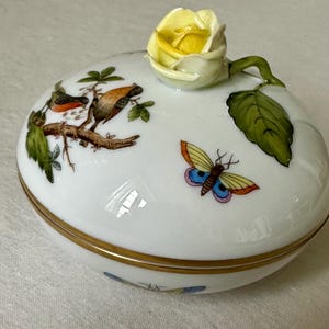May include: A white porcelain trinket box with a gold rim, featuring painted birds, a butterfly, and a yellow rose on the lid. The box has a glossy finish and a detailed floral design, perfect for storing small items.