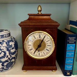 May include: A wooden mantel clock with a dark brown finish, featuring a gold-toned circular face with black Roman numerals and a central decorative gold medallion. The clock is topped with a small brass finial and rests on four small brass feet.