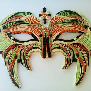 Könnte beinhalten: Eine schwarze, orange und grüne Papier-Quilling-Maske mit filigranen Details. Die Maske hat ein einzigartiges Design mit einem spitzen Kinn und einem dekorativen Oberteil.