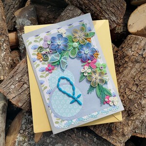 May include: A handmade greeting card with a blue, green, and yellow paper flower bouquet on a gray background. The card has a white border and a light blue circle with a beaded design in the center.