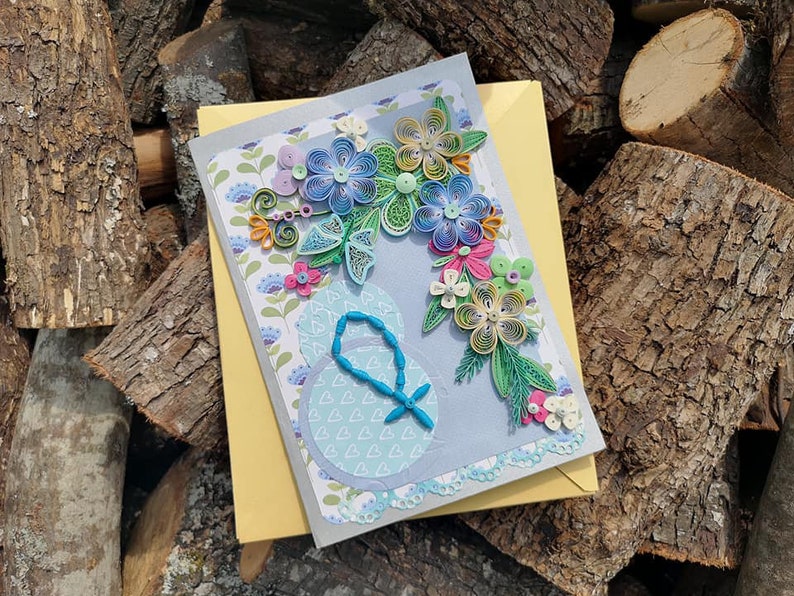 May include: A handmade greeting card with a blue, green, and yellow paper flower bouquet on a gray background. The card has a white border and a light blue circle with a beaded design in the center.
