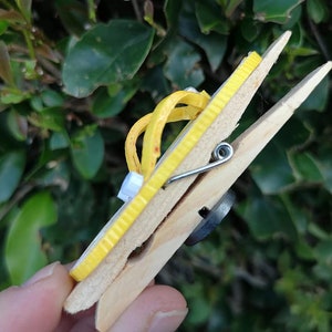 Op de afbeelding: Een gele houten wasknijper met een gele rubberen band eromheen gewikkeld. De wasknijper wordt vastgehouden door een hand.