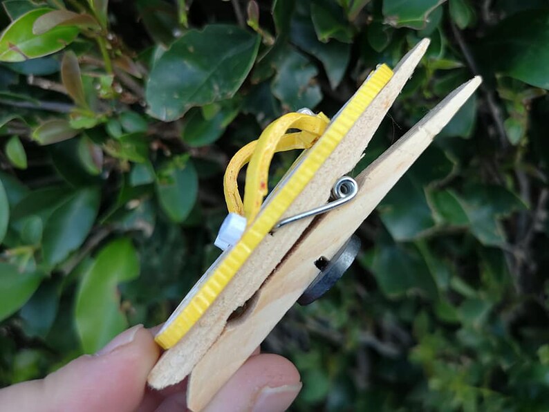 Op de afbeelding: Een gele houten wasknijper met een gele rubberen band eromheen gewikkeld. De wasknijper wordt vastgehouden door een hand.