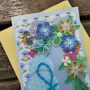 May include: A handmade greeting card with a blue background featuring a paper quilling design of flowers in shades of blue, green, yellow, and pink. The flowers are arranged in a bouquet shape and are surrounded by a border of hearts.
