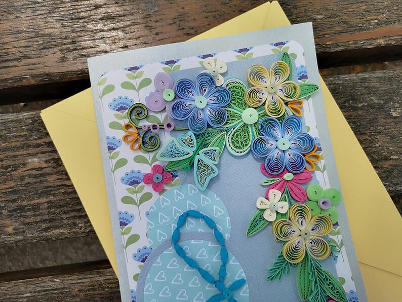 May include: A handmade greeting card with a blue background featuring a paper quilling design of flowers in shades of blue, green, yellow, and pink. The flowers are arranged in a bouquet shape and are surrounded by a border of hearts.