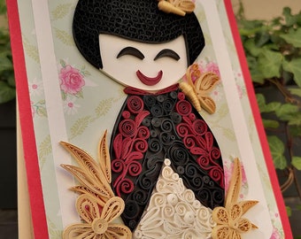 May include: A paper quilling art piece depicting a Japanese doll with black hair, red and black clothing, and white and gold accents. The doll is surrounded by flowers and butterflies.
