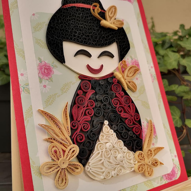 May include: A paper quilling art piece depicting a Japanese doll with black hair, red and black clothing, and white and gold accents. The doll is surrounded by flowers and butterflies.