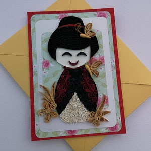 May include: A handmade greeting card featuring a paper quilling art piece of a Japanese doll with a red and black kimono, white face, and black hair. The doll is surrounded by gold paper quilling flowers and butterflies.