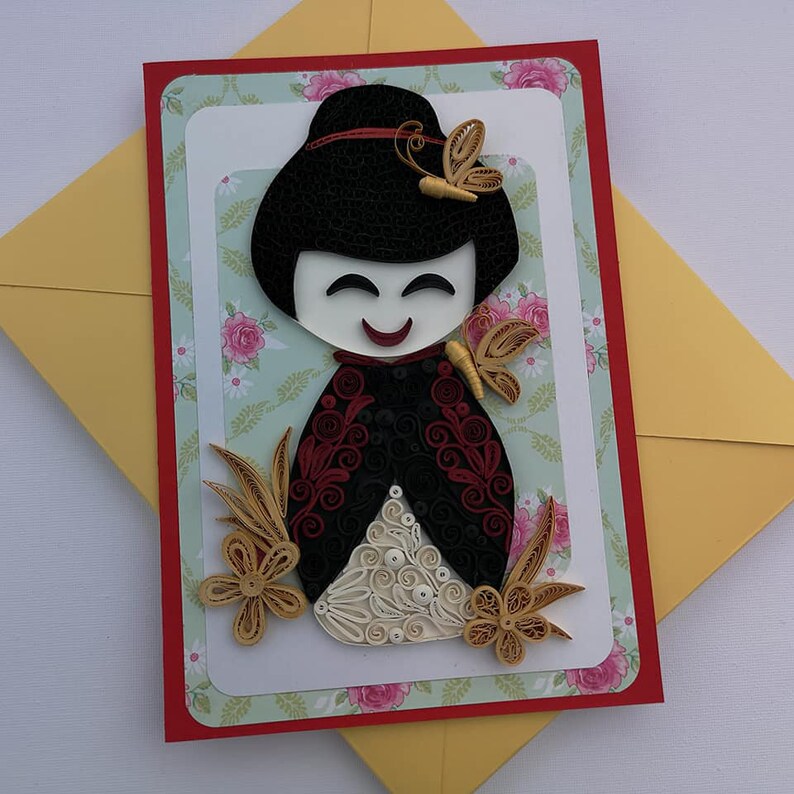 May include: A handmade greeting card featuring a paper quilling art piece of a Japanese doll with a red and black kimono, white face, and black hair. The doll is surrounded by gold paper quilling flowers and butterflies.