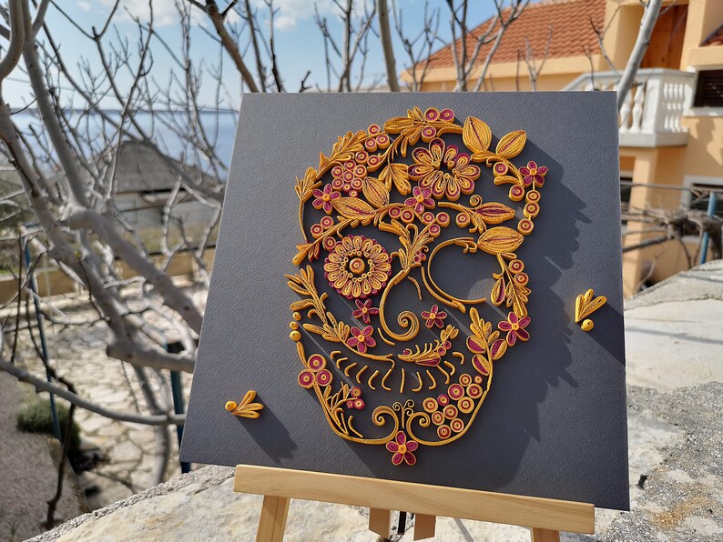 K&ouml;nnte beinhalten: Ein Papier-Quilling-Kunstwerk, das einen Sch&auml;del mit aufwendigen floralen Mustern in Gelb- und Burgunder-T&ouml;nen darstellt.